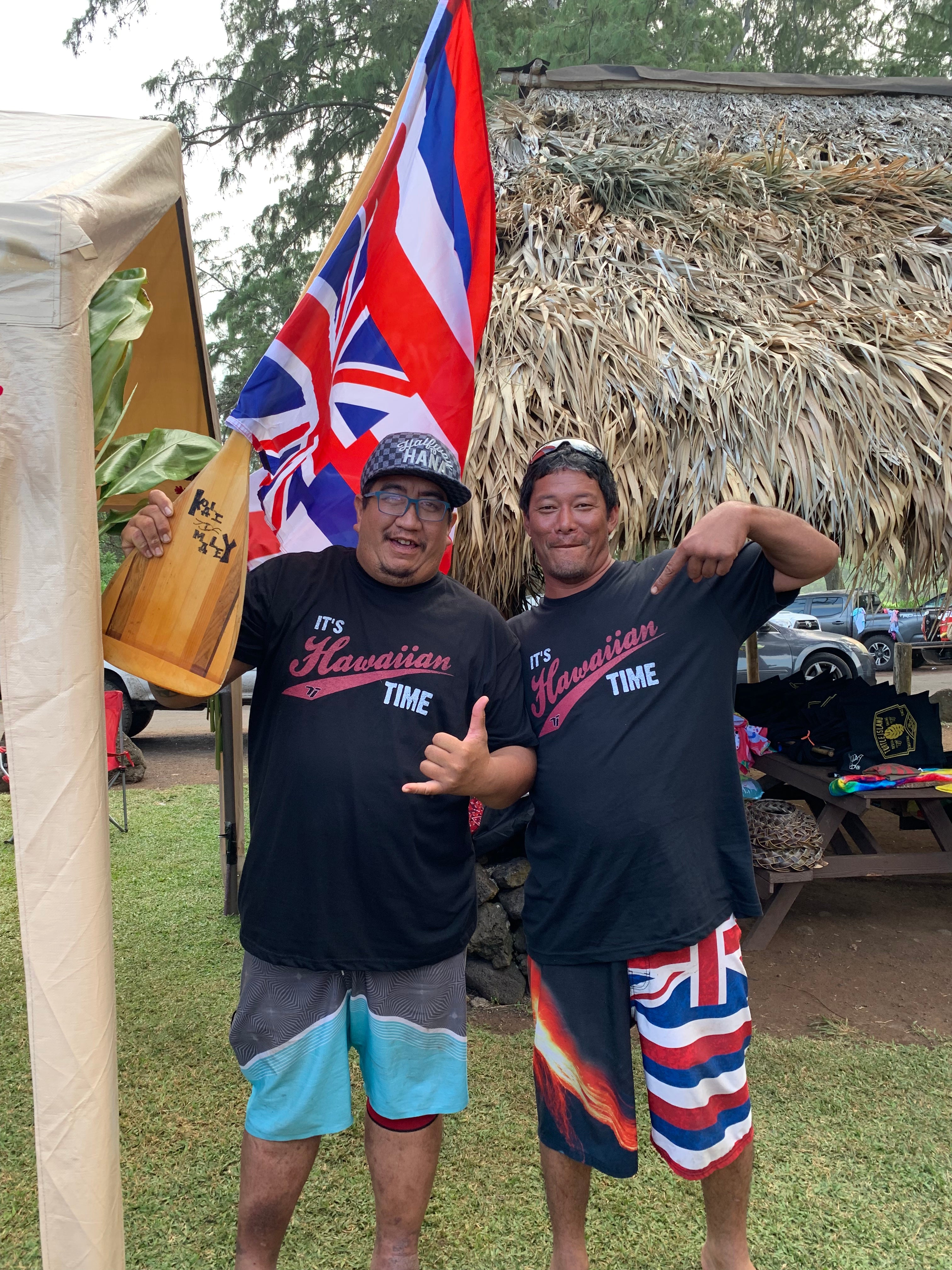 Two people wearing black shirts with visible text, standing outdoors with a flag and wooden object.